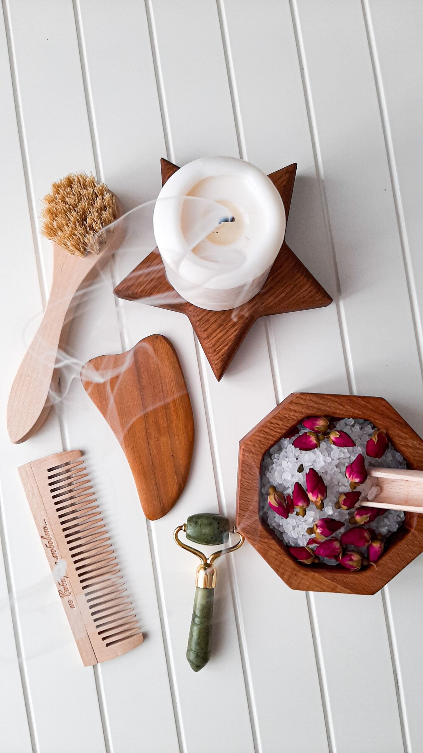 Flat lay of spa accessories including a candle, brush, comb, jade roller, and rose petals.